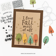 A handmade autumn-themed greeting card with the phrase 'I love FALL most of all' in brown lettering on a kraft paper background, surrounded by layered die-cut paper trees in green, yellow, and orange. The card is displayed on a white background alongside other crafting elements and a clear plastic die-cut template.
