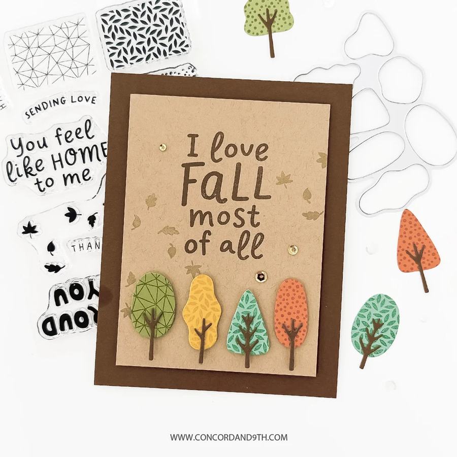A handmade autumn-themed greeting card with the phrase 'I love FALL most of all' in brown lettering on a kraft paper background, surrounded by layered die-cut paper trees in green, yellow, and orange. The card is displayed on a white background alongside other crafting elements and a clear plastic die-cut template.