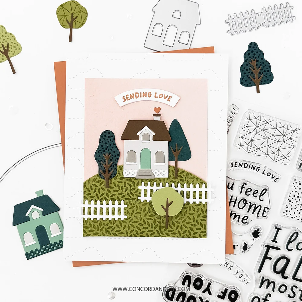 A handmade greeting card featuring a cute house illustration with the text 'SENDING LOVE' above it, styled on a white surface with craft cutouts and stamps around it.