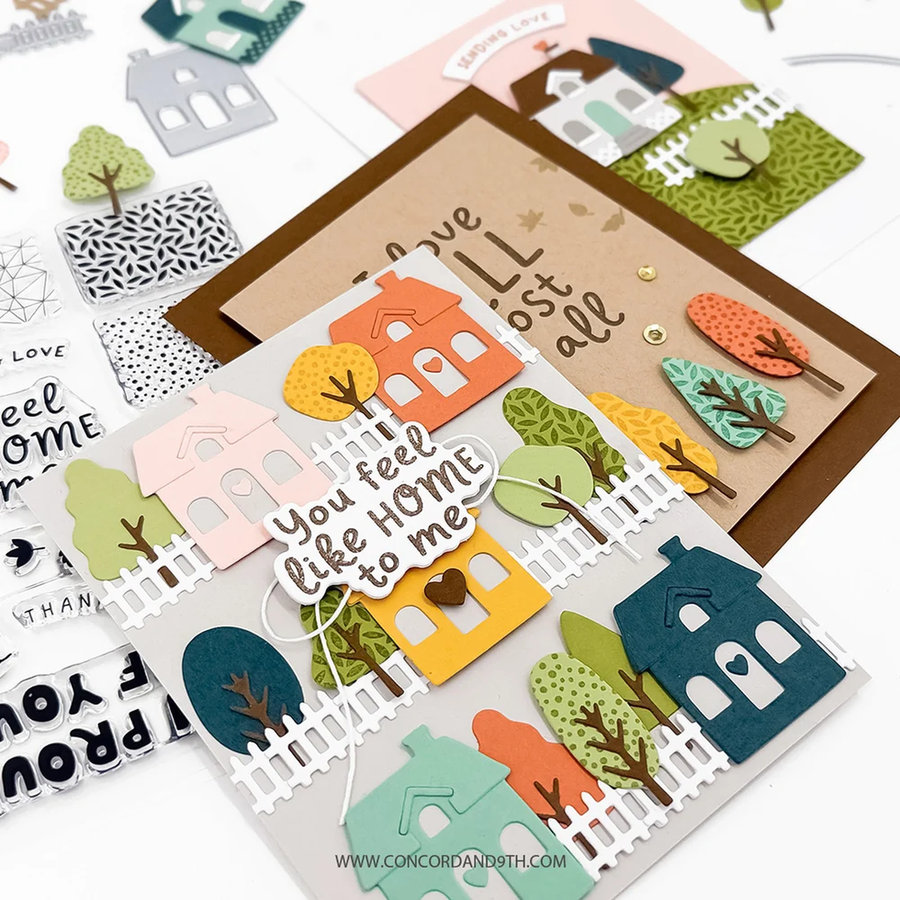 A handmade layered greeting card featuring autumn-themed houses and trees, displayed alongside other cards and embellishments on a white background. The main card has a kraft brown base with layered pastel and autumn-colored houses, white picket fences, and tree cutouts, with the phrase 'You feel like HOME to me' on a white script overlay.