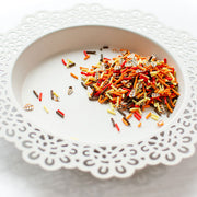 A pile of colorful clay confetti, including orange, red, yellow, brown, and white pieces, arranged on a white scalloped plate against a white background. Pretty Pink Posh Autumn Woods Clay Confetti