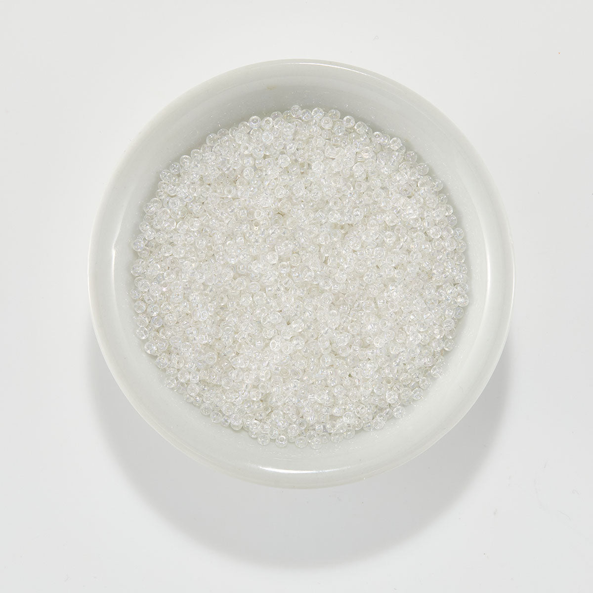 Clear seed beads filled with white glitter, displayed in a small white ceramic bowl on a clean white background. Spellbinders Clear Iridescent Beads