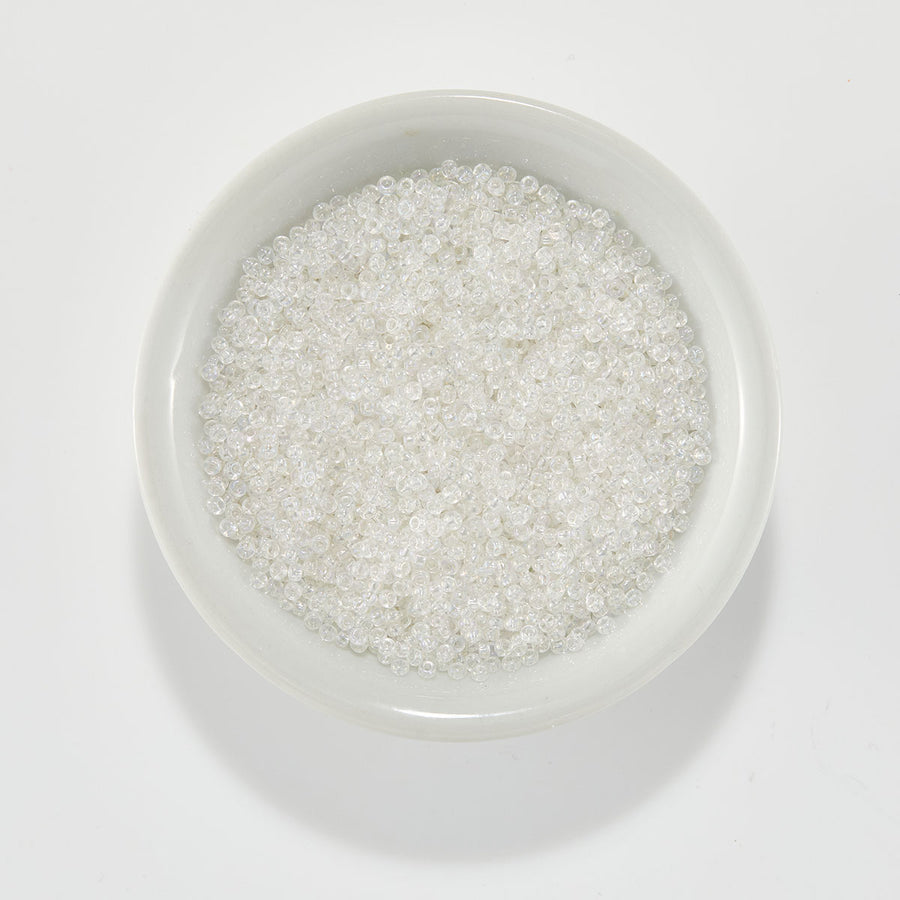 Clear seed beads filled with white glitter, displayed in a small white ceramic bowl on a clean white background. Spellbinders Clear Iridescent Beads
