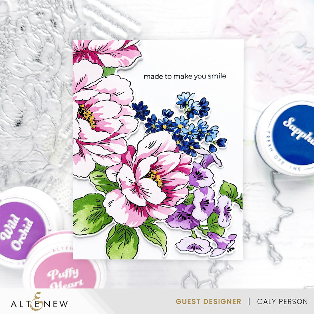 A floral card design displayed on a white surface, featuring large pink and purple flowers, blue blossoms, green leaves, and the printed phrase 'made to make you smile.' Surrounding the card are craft ink pads and embossed metal floral elements, with visible Altenew branding and guest designer text at the bottom.