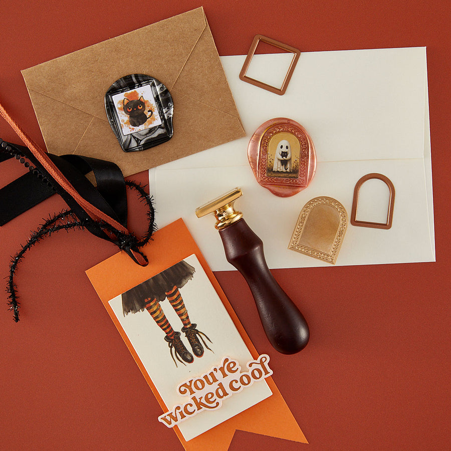 A flat-lay of a Halloween-themed handmade gift tag on a terracotta background, featuring a brown kraft envelope, a white card with a ghost illustration, an orange tag reading 'You're Wicked cool!', a dark brown leather seal stamp handle, gold metal frame pieces, and a round pink wax seal. Spellbinders 3D Photo & Floral Arch Frame Inserts Etched Dies