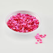 A white bowl filled with shiny hot pink sequins, with a few scattered on the white surface beside it.