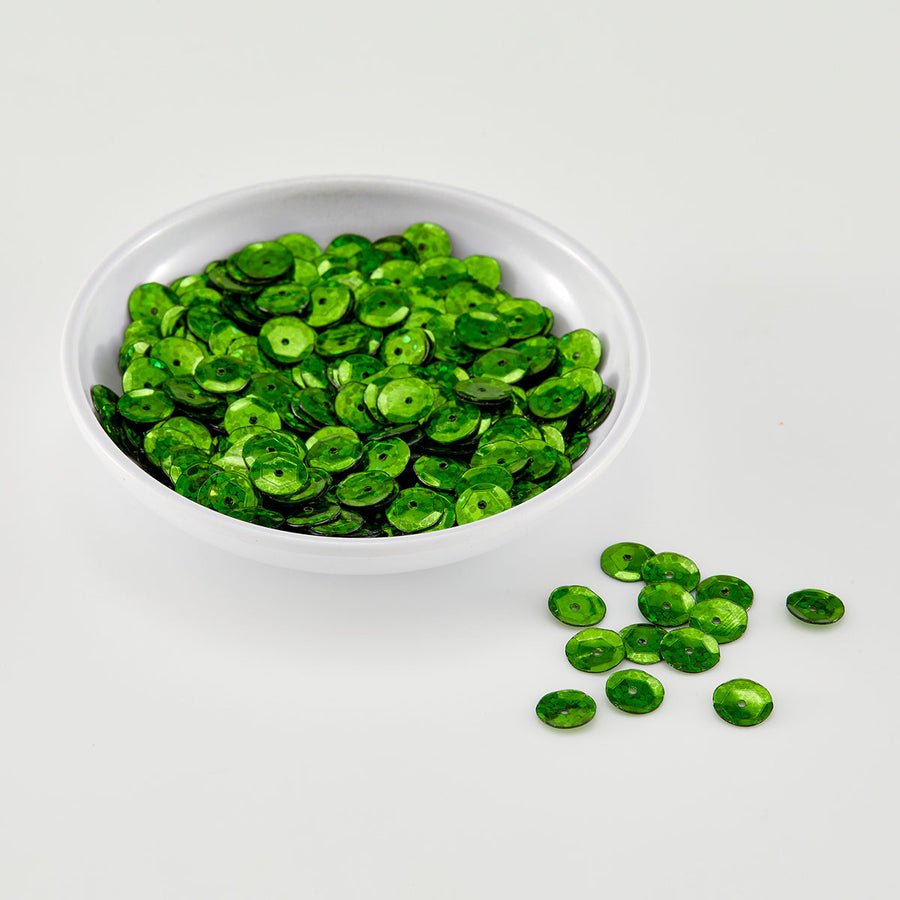 Green sequins in a small white bowl with a few scattered on a white surface.