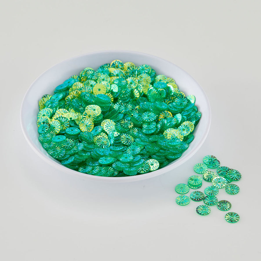 A white bowl filled with small round green sequins or embellishments, with a few scattered outside the bowl on a light gray background.