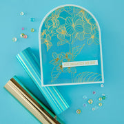 A flat-lay product shot of a handmade greeting card with a turquoise blue background, gold floral line art, and a white banner reading 'YOU BRIGHTEN MY DAY'. The card is styled with rolls of turquoise and gold metallic paper and scattered rhinestone embellishments on a solid turquoise blue background.
