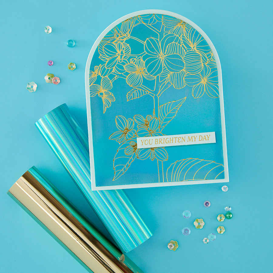 A flat-lay product shot of a handmade greeting card with a turquoise blue background, gold floral line art, and a white banner reading 'YOU BRIGHTEN MY DAY'. The card is styled with rolls of turquoise and gold metallic paper and scattered rhinestone embellishments on a solid turquoise blue background.