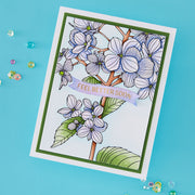 A handmade greeting card with a botanical illustration of white and lavender flowers, green leaves, and a lavender banner reading 'FEEL BETTER SOON!' in gold lettering, photographed on a bright blue background with small scattered decorative beads.