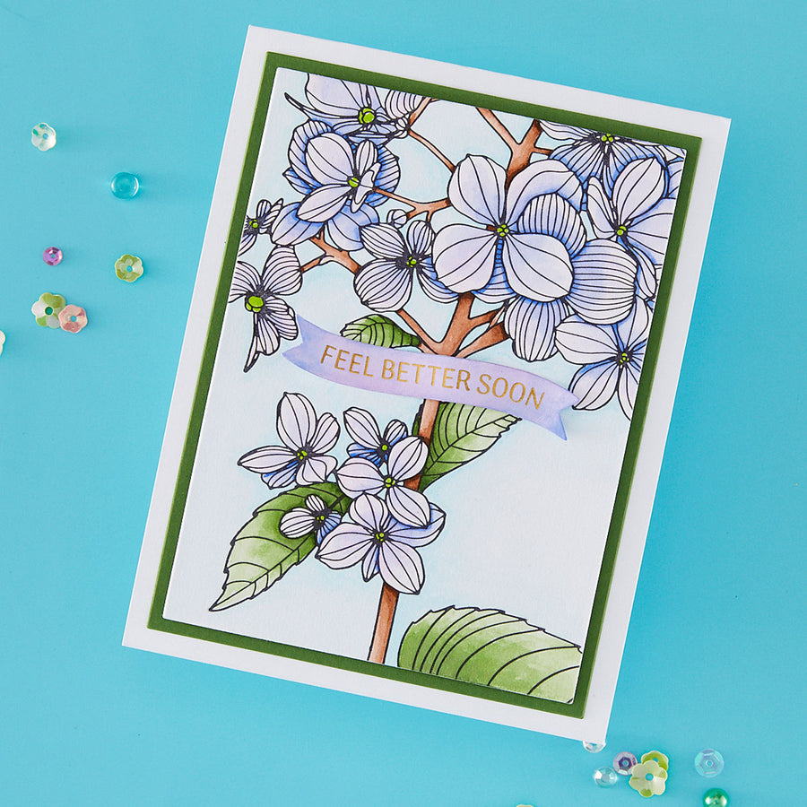 A handmade greeting card with a botanical illustration of white and lavender flowers, green leaves, and a lavender banner reading 'FEEL BETTER SOON!' in gold lettering, photographed on a bright blue background with small scattered decorative beads.