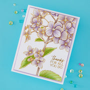 A handmade greeting card with a white background, featuring a detailed illustration of light purple hydrangea flowers with green leaves and gold outlines. The right side of the card has the printed text 'thanks FOR ALL YOU DO' in gold.