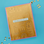A handmade greeting card with a gold foil embossed pattern of windows and a pink border, featuring a white banner that reads 'YOU BRIGHTEN MY DAY'. The card is photographed on a bright blue background with scattered gold sequins.