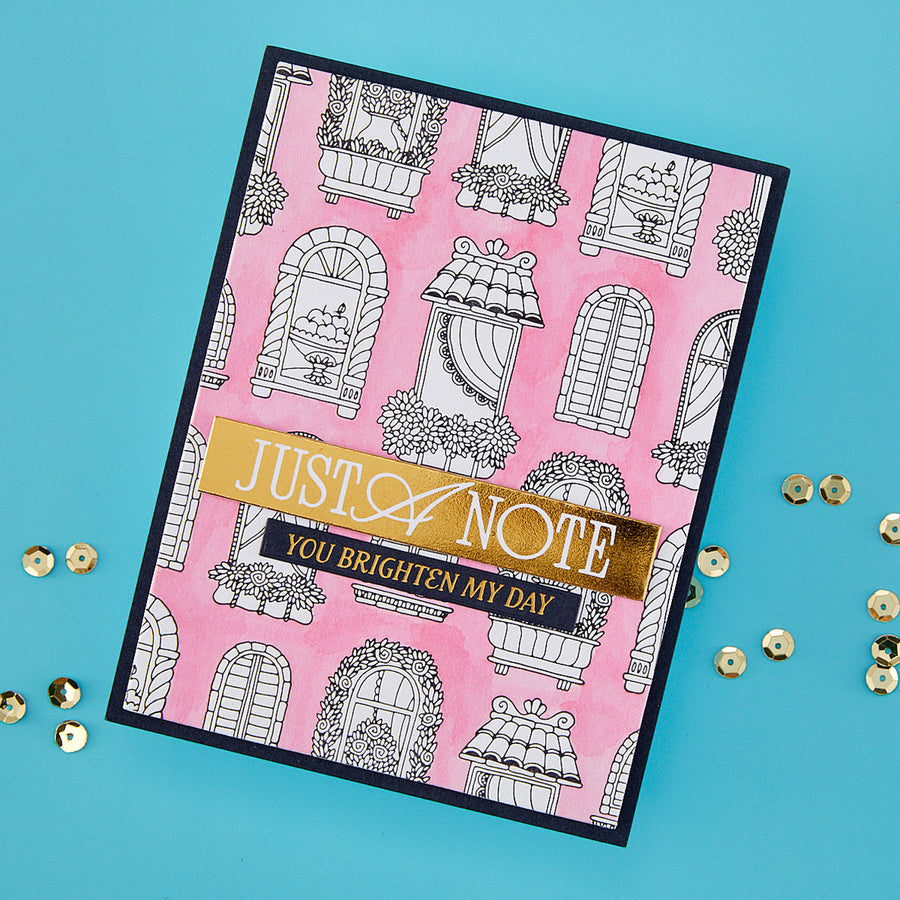 A handmade greeting card with a pink watercolor background, black line-art window illustrations, and a gold foil banner reading 'JUST A NOTE YOU BRIGHTEN MY DAY', photographed on a bright blue background with scattered gold sequins.