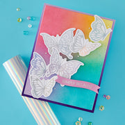 A handmade layered card featuring white die-cut butterfly silhouettes over a vibrant pink, orange, and teal gradient background, with a pink banner reading 'SIMPLY BECAUSE'. The card is styled on a bright blue background next to a roll of iridescent wrapping paper and scattered decorative beads.