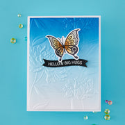 A handmade greeting card featuring a white embossed floral background, a blue-to-white gradient top section, a die-cut butterfly with orange, black, and white patterned wings, and a black banner reading 'HELLO & BIG HUGS'. The card is photographed on a bright turquoise background with scattered decorative rhinestones.