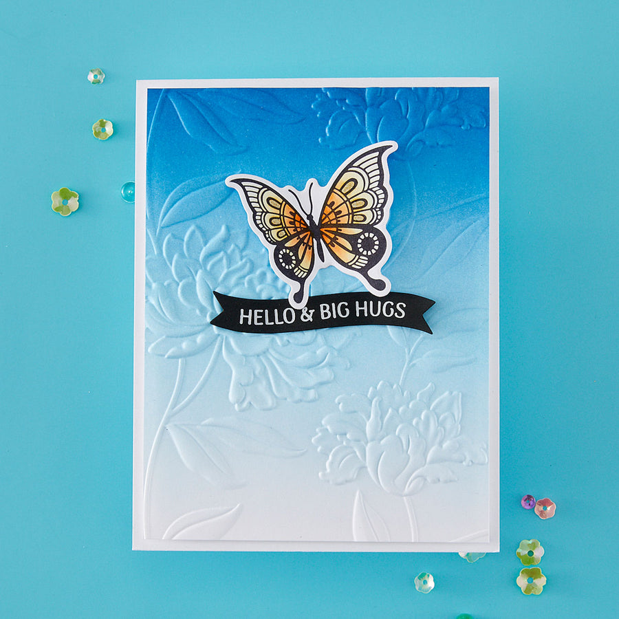 A handmade greeting card featuring a white embossed floral background, a blue-to-white gradient top section, a die-cut butterfly with orange, black, and white patterned wings, and a black banner reading 'HELLO & BIG HUGS'. The card is photographed on a bright turquoise background with scattered decorative rhinestones.