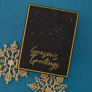 A square black holiday greeting card with a gold border, subtle snowflake pattern, and gold foil script reading 'Season's Greetings,' styled on a teal background with decorative gold snowflakes.