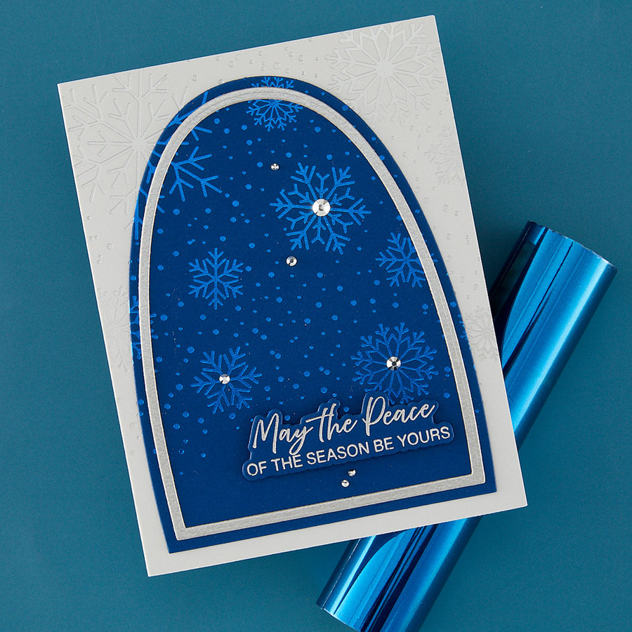 A handmade holiday greeting card with a blue arched panel featuring snowflakes, dots, and the text 'May the Peace OF THE SEASON BE YOURS,' shown on a teal background with metallic blue wrapping paper.