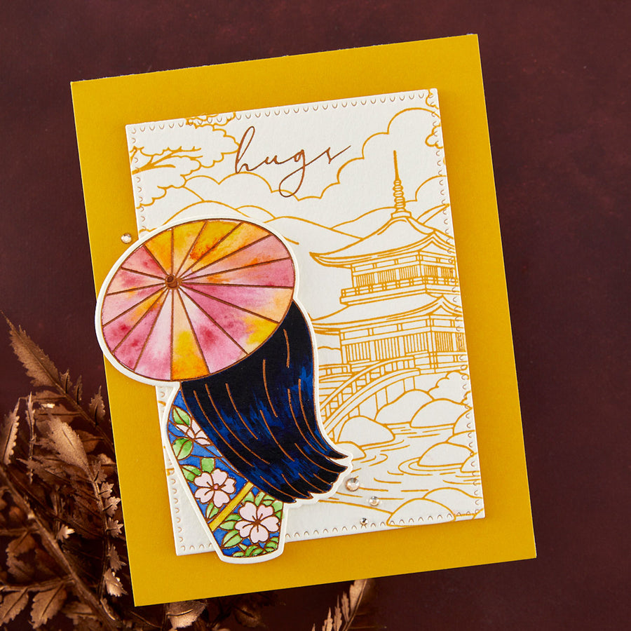 A handmade greeting card featuring a Japanese-inspired design with a watercolor umbrella, dark blue hair, and gold line art of a pagoda and bridge, displayed on a yellow cardstock over a dark brown background with dried foliage.