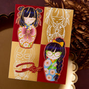 A decorative card or art print featuring two stylized doll illustrations in a red-and-gold four-panel layout, photographed on a reflective gold surface with dried botanicals in the background.