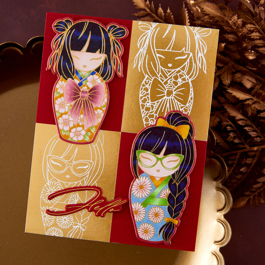 A decorative card or art print featuring two stylized doll illustrations in a red-and-gold four-panel layout, photographed on a reflective gold surface with dried botanicals in the background.