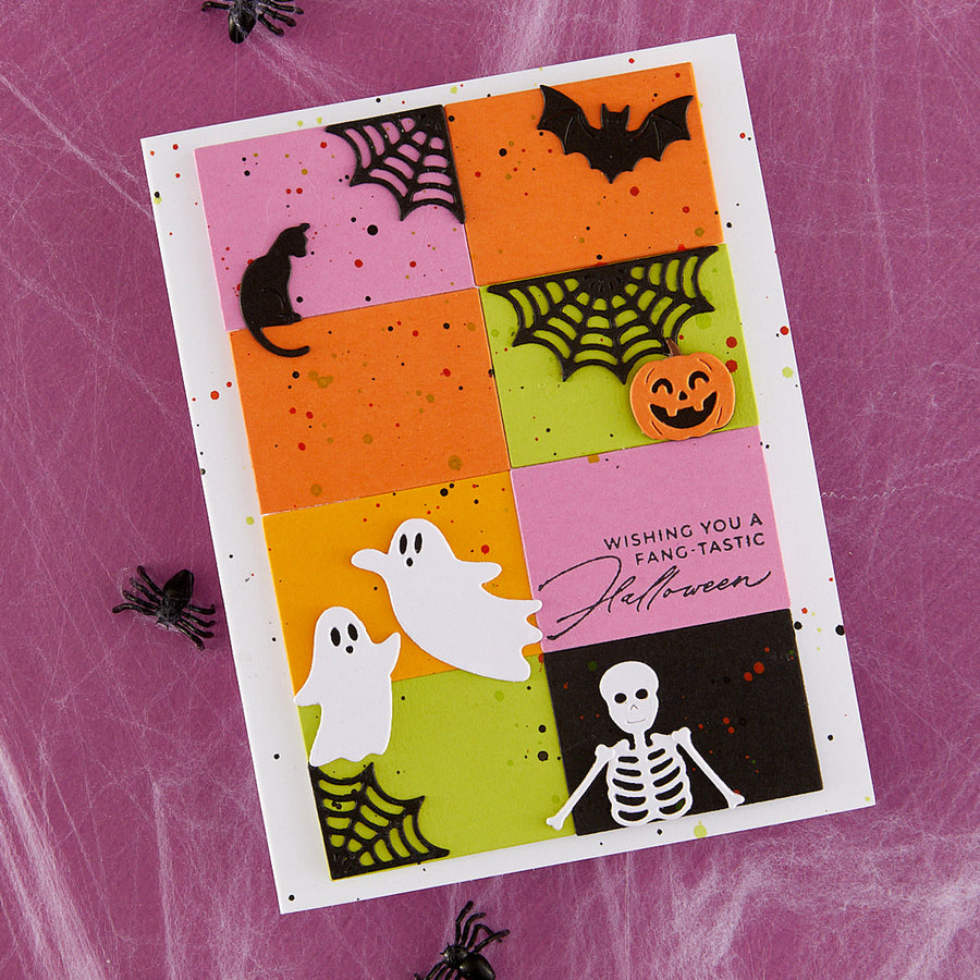 A handmade Halloween greeting card with a colorful grid design featuring a black cat, bat, spiderwebs, jack-o'-lantern, ghosts, skeleton, and the phrase 'Wishing You A Fang-Tastic Halloween,' photographed on a pink spiderweb-patterned background with small black spider props.