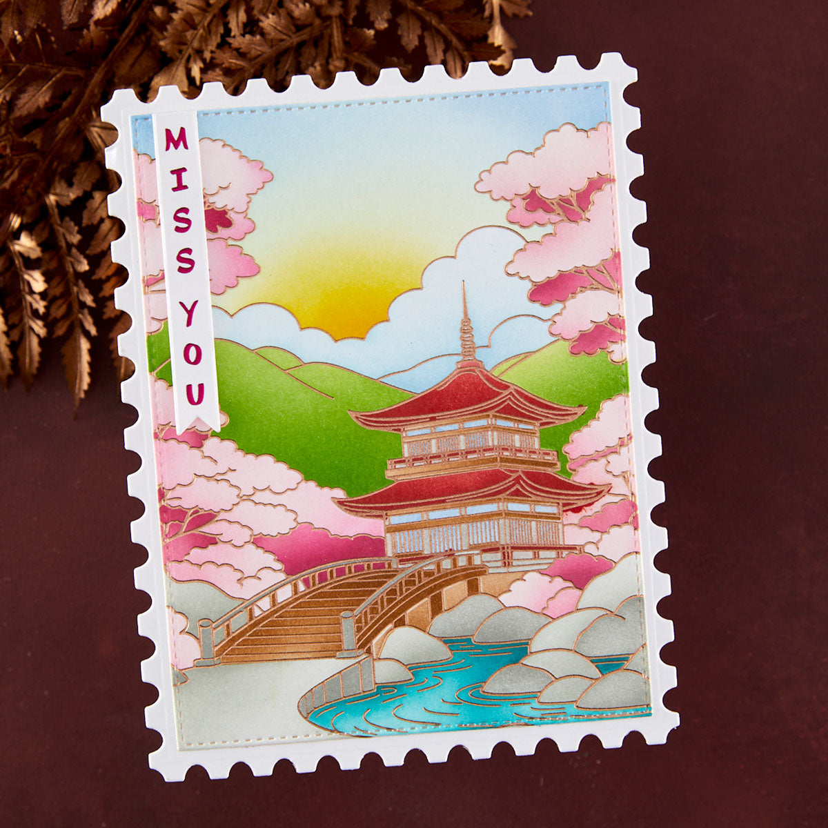A postage stamp-style handmade greeting card with a Japanese pagoda, bridge, river, mountains, and pink cherry blossoms, plus the text 'MISS YOU' on the left side, photographed on a dark brown surface with decorative brown leaves in the corner.