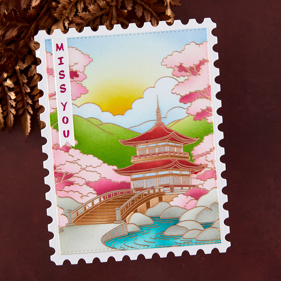 A postage stamp-style handmade greeting card with a Japanese pagoda, bridge, river, mountains, and pink cherry blossoms, plus the text 'MISS YOU' on the left side, photographed on a dark brown surface with decorative brown leaves in the corner.