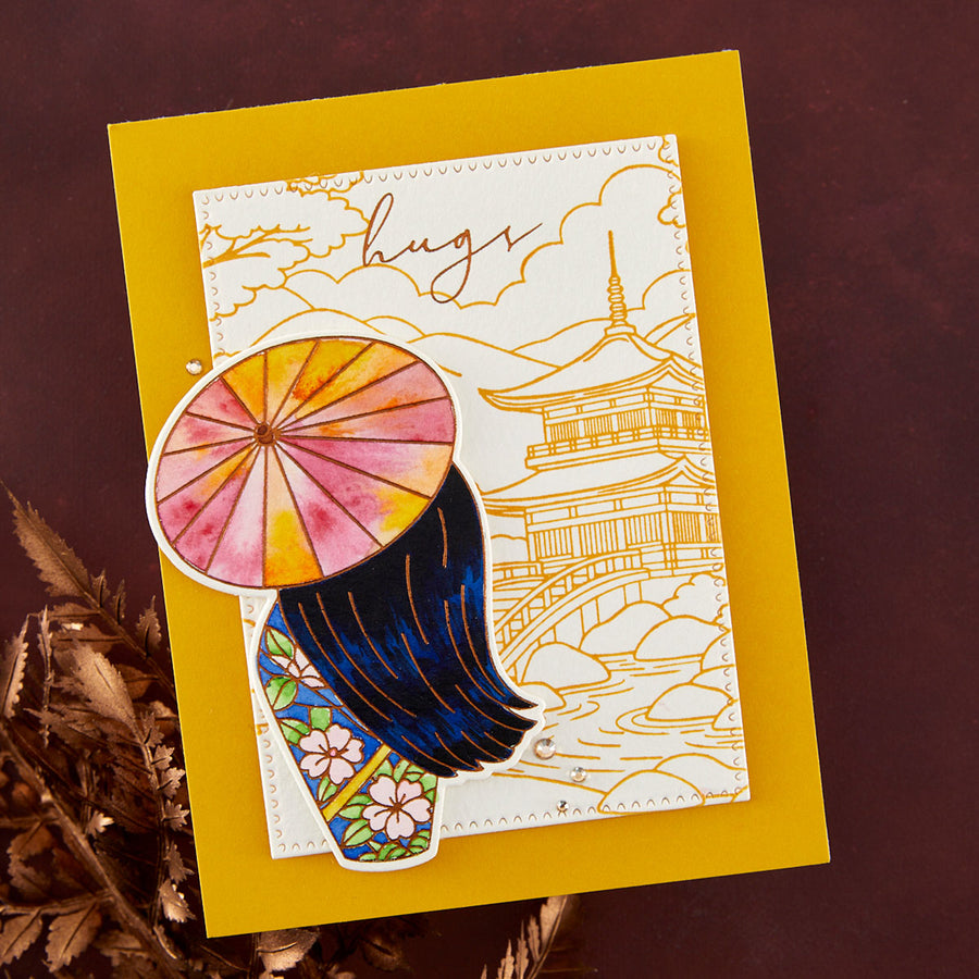 A handmade layered greeting card featuring a Japanese-inspired design with a watercolor umbrella, dark blue hair, floral pattern, and gold line art of a pagoda and bridge, placed on a yellow cardstock over a dark brown background with dried leaves.