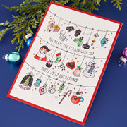 A handmade Christmas greeting card with a red border, featuring hand-drawn holiday ornaments and illustrations including a Santa Claus, snowman, teddy bear, candy cane, and hot chocolate. The card is displayed on a dark blue surface with green pine branches and colorful Christmas ornaments as props. Spellbinders Decorate the Season Registration Press + Foil Plate