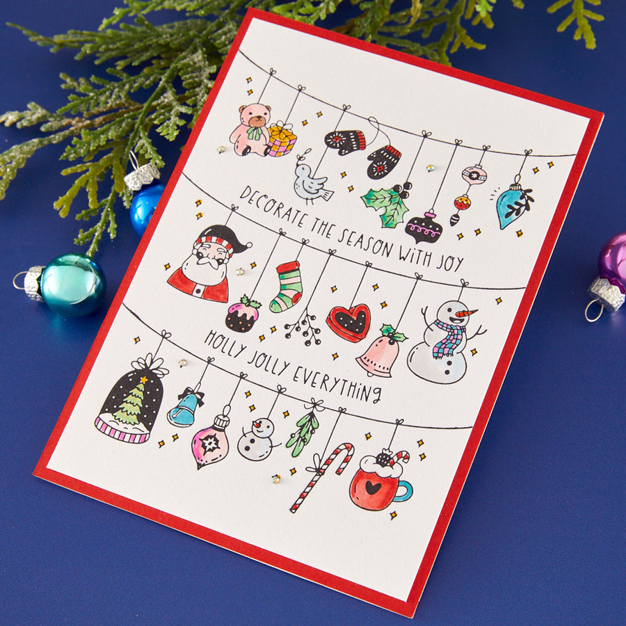 A handmade Christmas greeting card with a red border, featuring hand-drawn holiday ornaments and illustrations including a Santa Claus, snowman, teddy bear, candy cane, and hot chocolate. The card is displayed on a dark blue surface with green pine branches and colorful Christmas ornaments as props. Spellbinders Decorate the Season Registration Press + Foil Plate