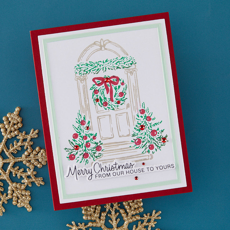 A handmade Christmas greeting card with a red border, featuring an illustrated front door decorated with a green wreath and red berries, placed on a teal background with gold decorative snowflakes.