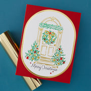 A handmade red Christmas greeting card with a white oval center featuring an illustrated holiday door with green wreaths and gold trim, plus the words 'Merry Christmas' in script. The card is photographed on a blue background with gold cylindrical props partially visible underneath.