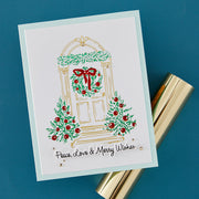 A handmade white greeting card with a holiday door illustration, green wreaths and garlands, red berries, and the text 'Peace, Love & Merry Wishes,' photographed on a blue background with a gold cylindrical prop beside it.