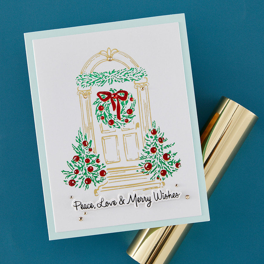 A handmade white greeting card with a holiday door illustration, green wreaths and garlands, red berries, and the text 'Peace, Love & Merry Wishes,' photographed on a blue background with a gold cylindrical prop beside it.