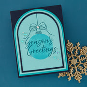 A handmade holiday greeting card with an arched layered design in teal, black, and light blue, featuring the words 'Season's Greetings' over a blue ornament graphic. The card is styled on a dark teal background next to a gold glitter snowflake.