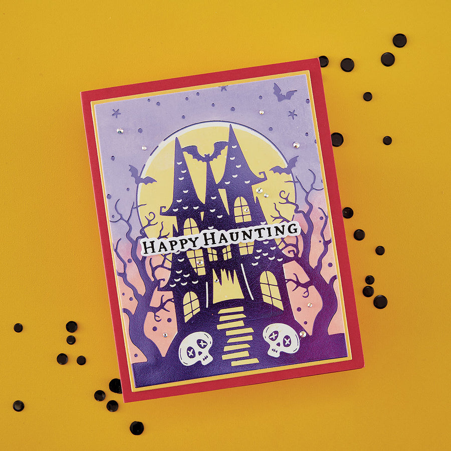 A Halloween-themed handmade greeting card featuring a haunted house, bats, skulls, and the words 'Happy Haunting' on a yellow background with scattered black confetti. Using Spellbinders BetterPress Society August 2026 (Haunted Hollow Press Plate & Die Set + Add-on Stencil Bundle)