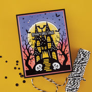 A Halloween handmade greeting card featuring a spooky haunted house, bats, stars, and the words 'Spooky Greetings' on a yellow background, styled with a roll of black-and-white twine and scattered black pom-poms. Using Spellbinders BetterPress Society August 2026 (Haunted Hollow Press Plate & Die Set + Add-on Stencil Bundle)