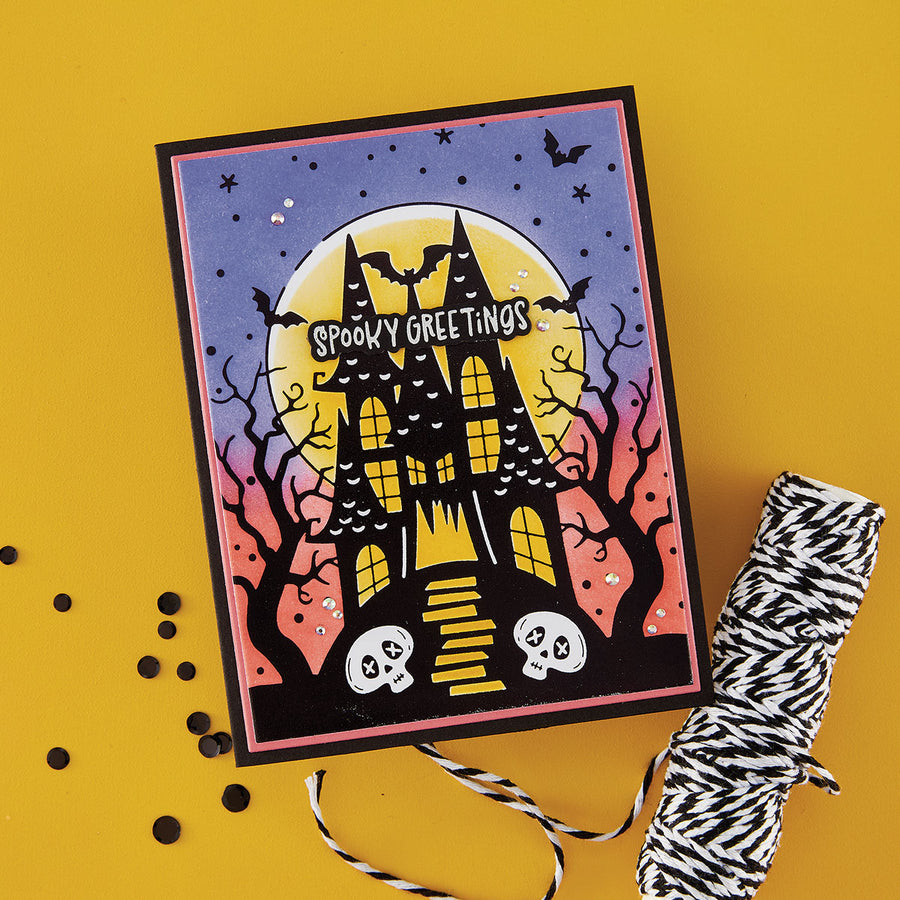 A Halloween handmade greeting card featuring a spooky haunted house, bats, stars, and the words 'Spooky Greetings' on a yellow background, styled with a roll of black-and-white twine and scattered black pom-poms. Using Spellbinders BetterPress Society August 2026 (Haunted Hollow Press Plate & Die Set + Add-on Stencil Bundle)