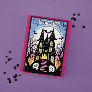 A Halloween handmade greeting card with a spooky haunted house illustration, placed on a purple background with scattered black confetti. The card has a bright pink border and the text 'SPOOKY GREETINGS' at the top. Using Spellbinders BetterPress Society August 2026 (Haunted Hollow Press Plate & Die Set + Add-on Stencil Bundle)