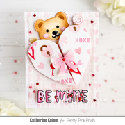 A handmade Valentine's Day card featuring a 3D teddy bear peeking behind a split heart design with the words 'XOXO' and 'BE MINE' in pink and red. The card is displayed on a white wooden surface with decorative flowers and confetti.