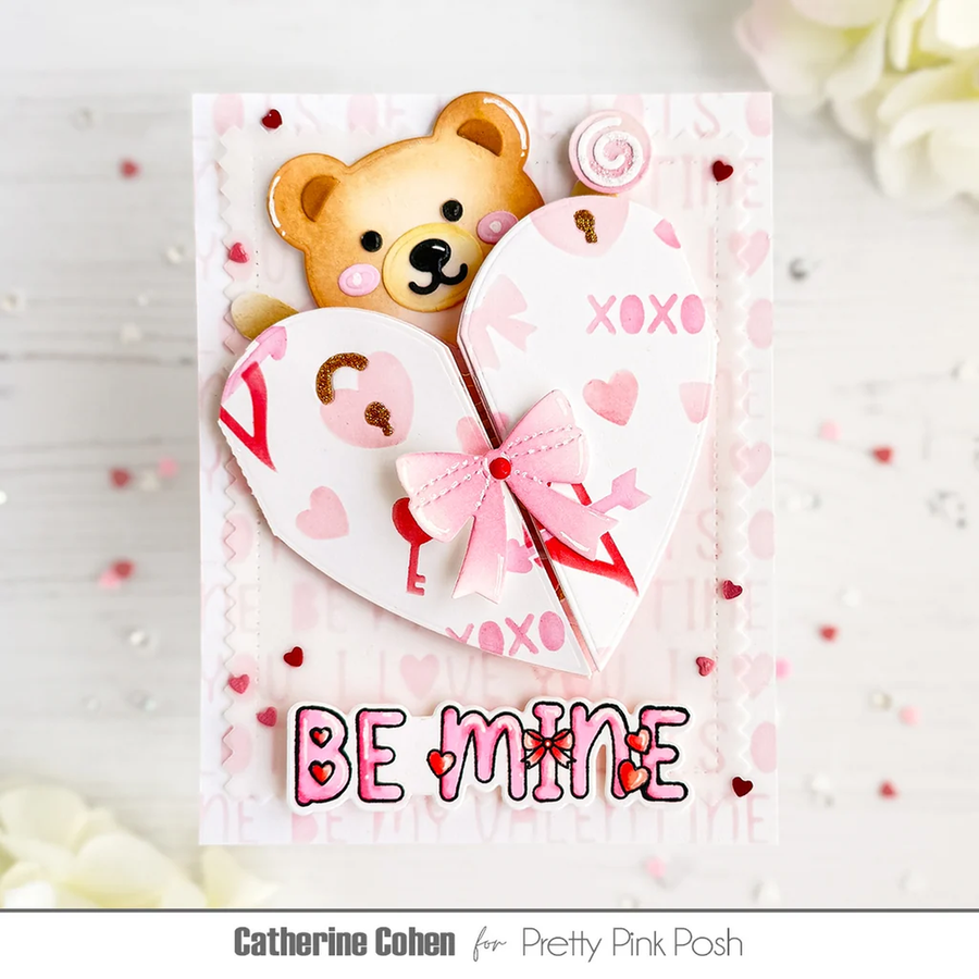 A handmade Valentine's Day card featuring a 3D teddy bear peeking behind a split heart design with the words 'XOXO' and 'BE MINE' in pink and red. The card is displayed on a white wooden surface with decorative flowers and confetti.