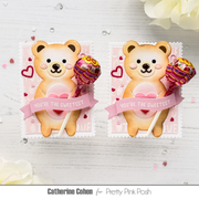 Two handmade Valentine's Day lollipops styled as teddy bears on pink stamp-like backing cards, displayed on a white wood surface with decorative flowers and small scattered hearts. The lollipops have multicolored striped candy designs and pink ribbon banners reading 'YOU'RE THE SWEETEST.