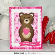 A handmade Valentine's Day greeting card featuring a brown bear holding a pink heart, set against a background of pink hearts and the words 'LOVE' and 'TIME'. The card is displayed on a reflective surface with decorative props, including a red 'Love' sculpture and white flowers.