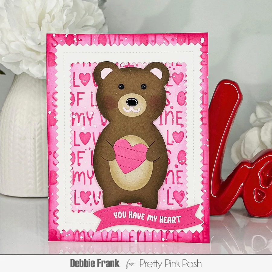 A handmade Valentine's Day greeting card featuring a brown bear holding a pink heart, set against a background of pink hearts and the words 'LOVE' and 'TIME'. The card is displayed on a reflective surface with decorative props, including a red 'Love' sculpture and white flowers.
