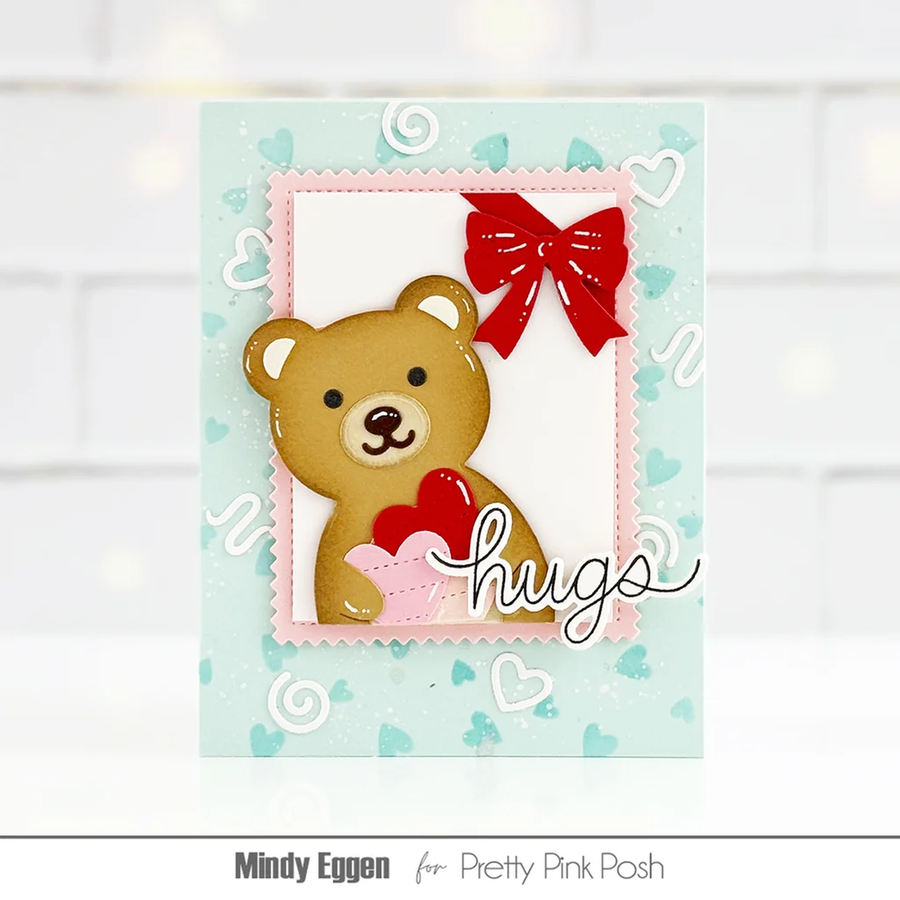 A handmade greeting card featuring a 3D brown teddy bear holding a red heart, framed by a pink scalloped border with a red bow on a light blue heart-patterned background. The card is signed 'Mindy Eggen for Pretty Pink Posh'.