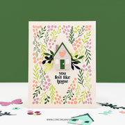 A handmade greeting card featuring a small house with a pink heart on its door, surrounded by layered green leaves and pink, orange, and purple flowers. The card reads 'you feel like home' in black text.