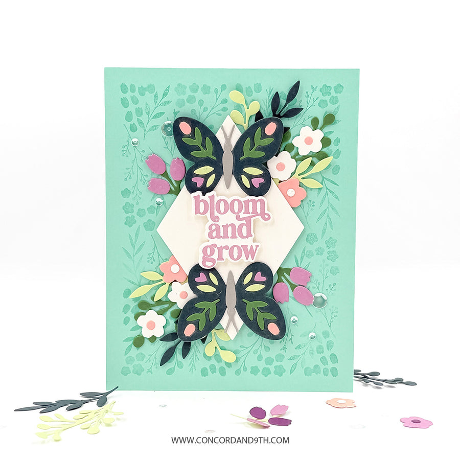 A handmade layered card featuring two black paper butterflies with green and pink details, surrounded by green leaves, pink and white flowers, and a central cream diamond with the words 'bloom and grow' in pink. The card is set against a white background with a few decorative paper petals scattered at the bottom.